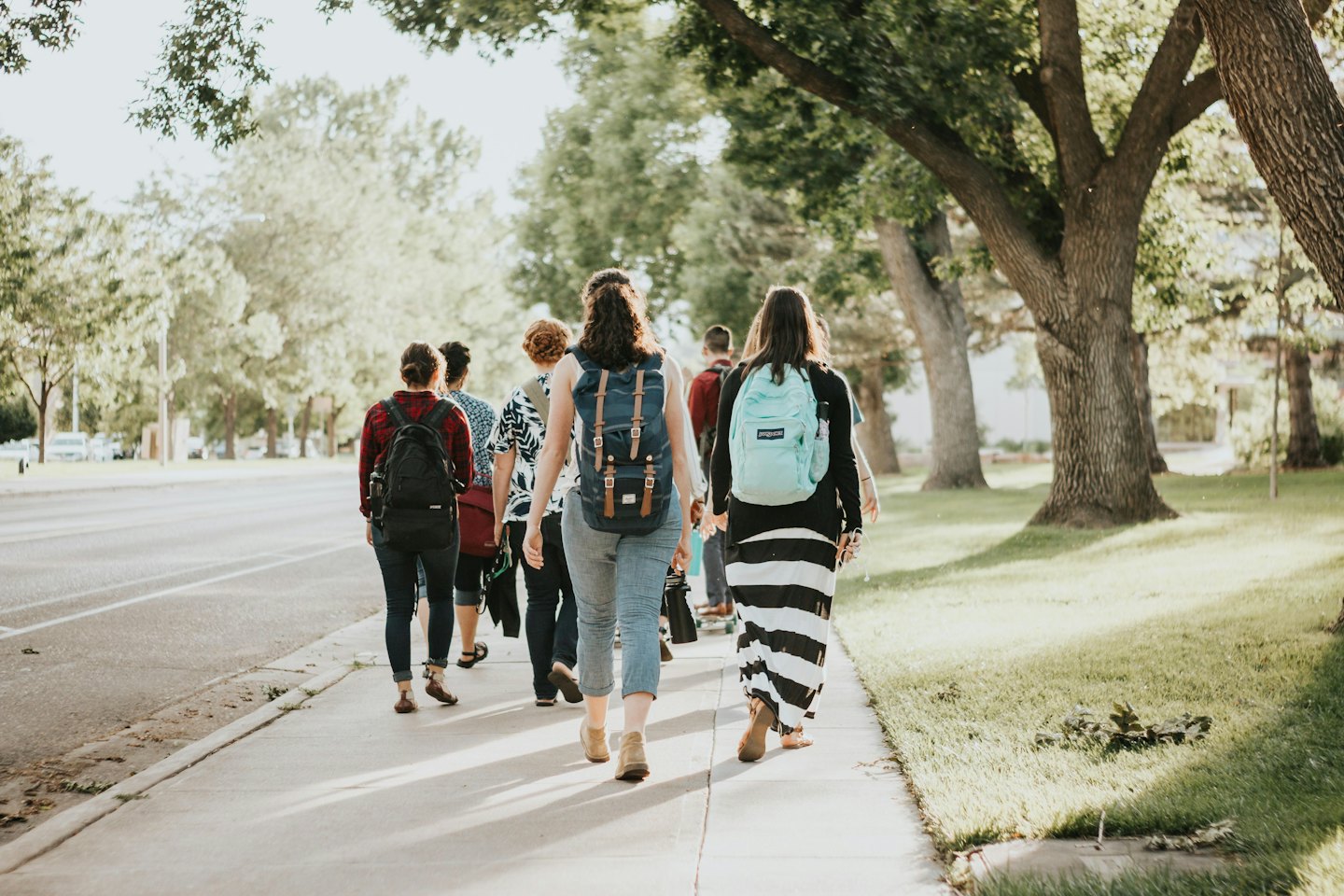 Diverse young people walk down a sunny sidewalk with backpacks.